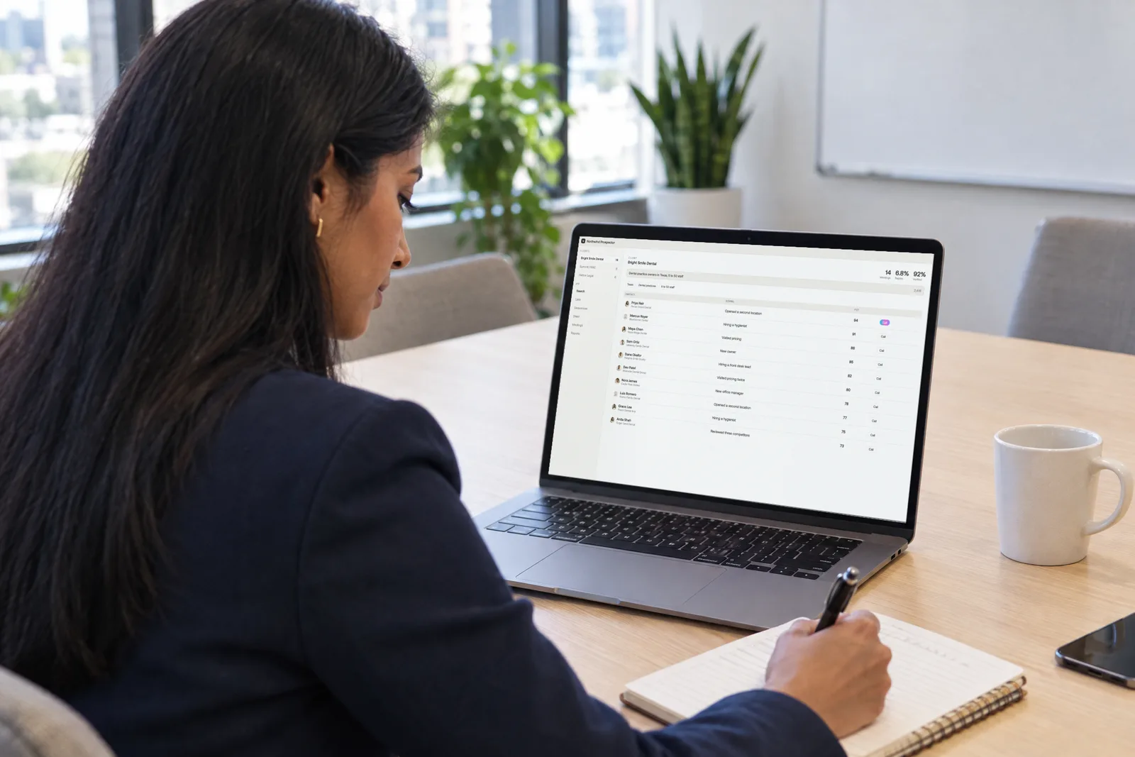 Seen over her shoulder, an account executive takes notes as she prepares for a call, her laptop open to her list of prospects