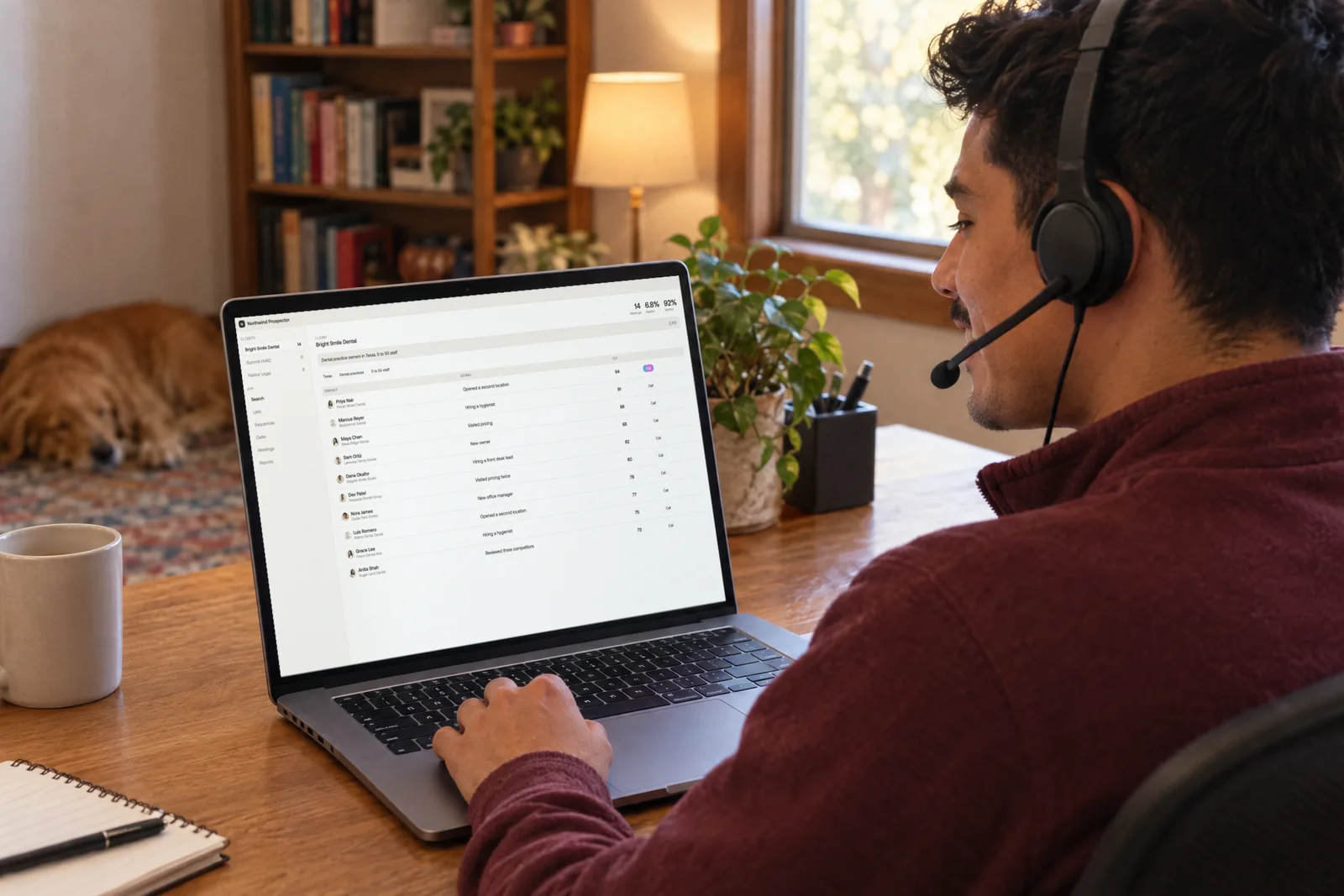 Seen over his shoulder, an SDR in a headset smiles on a call with his hand on the trackpad, his prospect list open and a dog asleep behind him